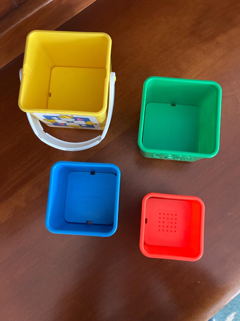 May include: Four brightly coloured stacking toy blocks. The largest is yellow with a white handle, the next is green, followed by blue, and the smallest is red with holes. The toys are square and designed for children.