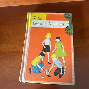 Peut inclure: Un livre relié vintage intitulé The New Learning Numbers 4 avec une couverture orange illustrée de quatre enfants jouant avec des anneaux sur un piquet. Le livre présente une bande jaune en haut et un motif de feuilles vertes sur la droite.