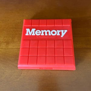 May include: A bright red plastic game case with a grid pattern on the surface. The word Memory is printed in large white letters across the top, with the text BY MILTON BRADLEY printed in smaller white letters directly underneath the main title.