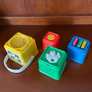 May include: A selection of vintage baby toys in bright colours. Includes a yellow cube with a handle, a red cube, a green cube with a white bird design, and a blue cube with three coloured cylinders. The toys are made of plastic.