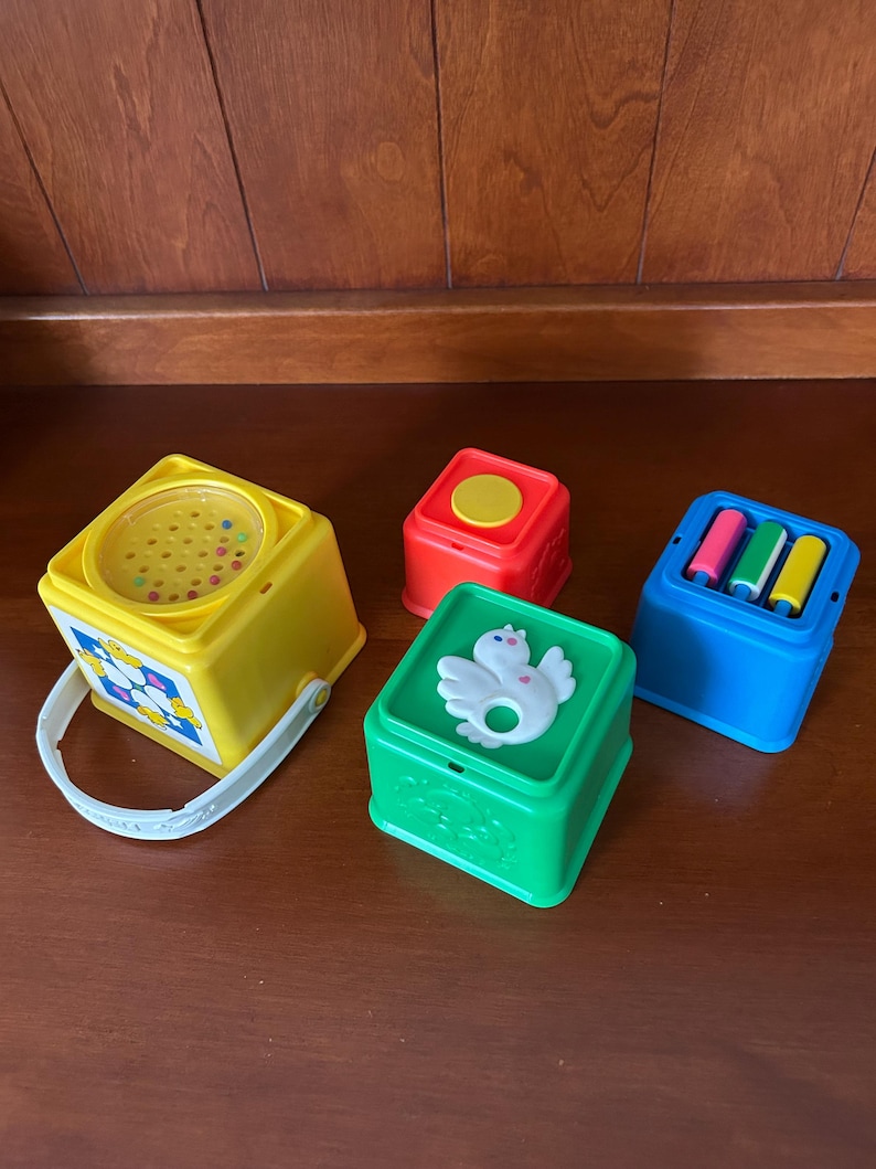 May include: A selection of vintage baby toys in bright colours. Includes a yellow cube with a handle, a red cube, a green cube with a white bird design, and a blue cube with three coloured cylinders. The toys are made of plastic.