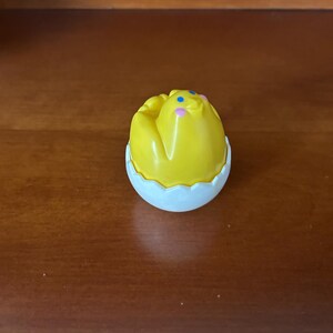 May include: A yellow chick toy with blue and pink accents, emerging from a white eggshell. The toy is likely a children's plaything, possibly a collectible or part of a larger set. The eggshell has a scalloped edge.