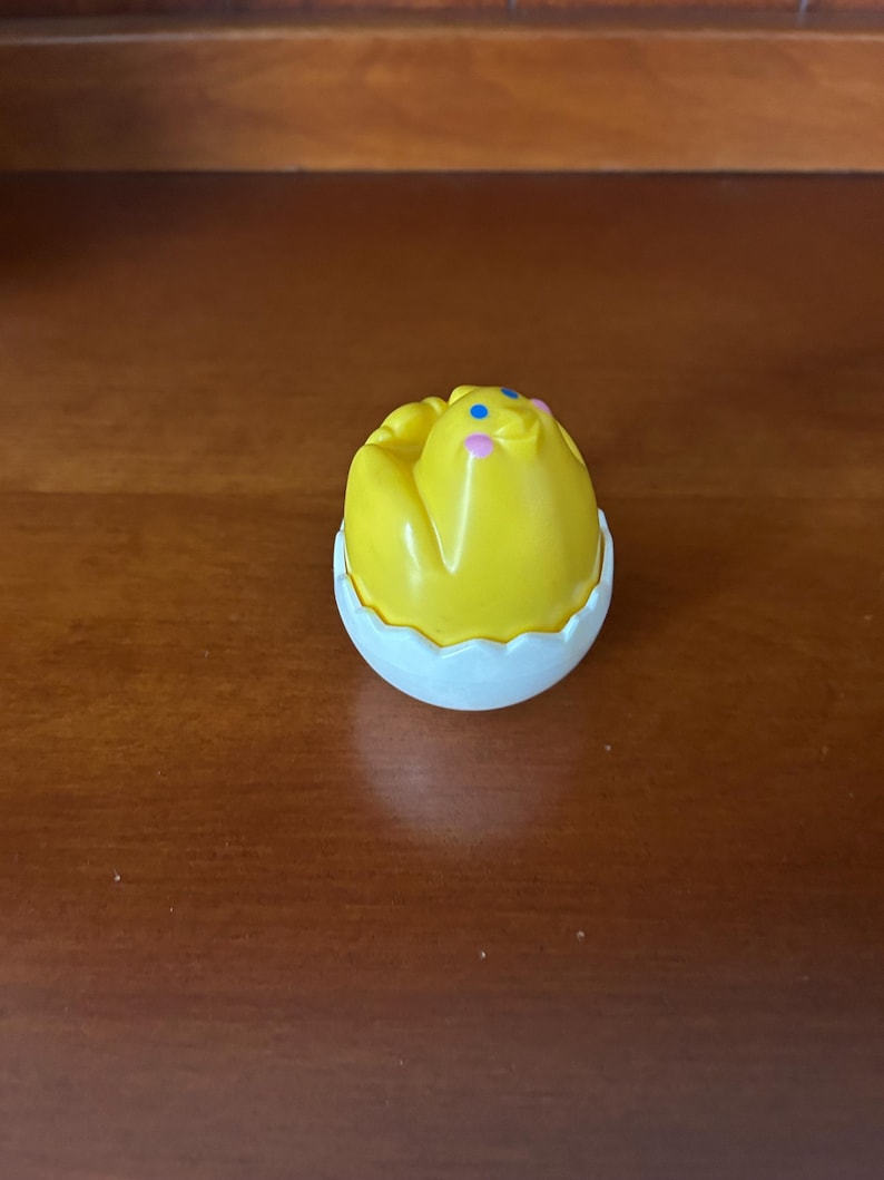 May include: A yellow chick toy with blue and pink accents, emerging from a white eggshell. The toy is likely a children's plaything, possibly a collectible or part of a larger set. The eggshell has a scalloped edge.