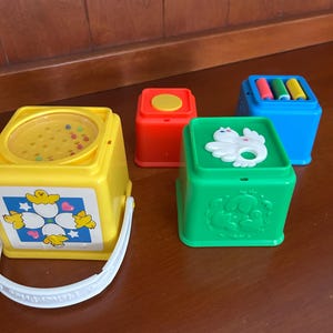 May include: A set of vintage baby toys in various colours. Includes a yellow bucket with a handle and a lid with small beads, a red block with a yellow circle, a green block with a white animal, and a blue block with colourful cylinders.