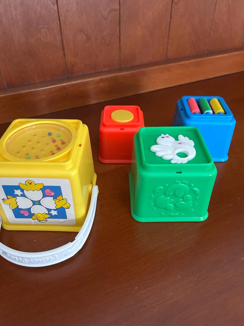 May include: A set of vintage baby toys in various colours. Includes a yellow bucket with a handle and a lid with small beads, a red block with a yellow circle, a green block with a white animal, and a blue block with colourful cylinders.