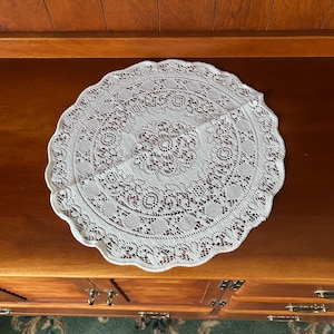 Könnte beinhalten: Ein rundes, weißes Spitzendeckchen mit einem komplizierten Blumen- und geometrischen Muster sowie einem gewellten Rand. Das Textilstück zeigt ein zentrales Medaillon-Design, das von konzentrischen Bändern aus Lochstickerei und feinen Fadendetails umgeben ist.
