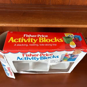May include: A vintage Fisher-Price Activity Blocks box. The box is red with white and blue accents and features the product name in large, colourful letters. The box is open, revealing the empty compartments for the blocks. The box also shows images of the blocks.