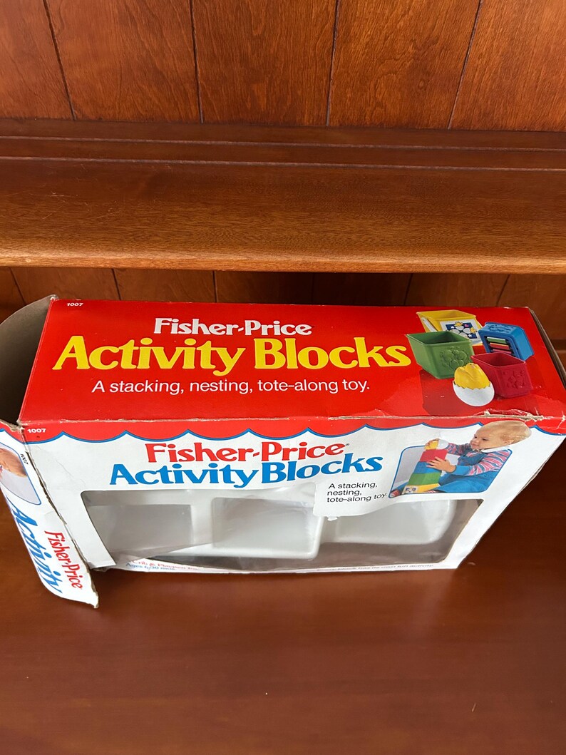 May include: A vintage Fisher-Price Activity Blocks box. The box is red with white and blue accents and features the product name in large, colourful letters. The box is open, revealing the empty compartments for the blocks. The box also shows images of the blocks.