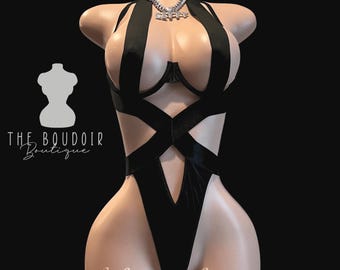 May include: Black lingerie with a criss-cross design and a plunging neckline. The lingerie features a velvet fabric and a delicate lace trim. The model is wearing a silver chain necklace with a pendant.