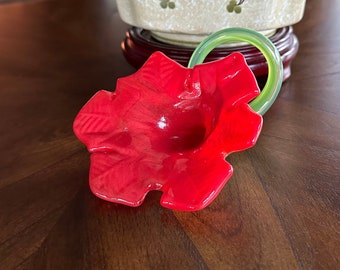 May include: A red glass flower with a green glass stem. The flower has a ruffled edge and a center hole. The stem is curled into a loop.