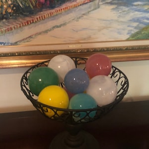 May include: A black metal bowl with a decorative stand holds a collection of colorful glass balls. The balls are in shades of green, blue, yellow, pink, and white.