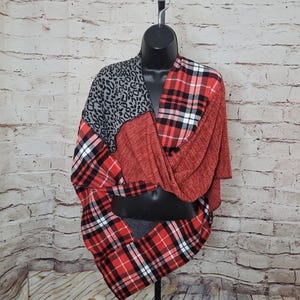 May include: A red, black, and white plaid top with a leopard print accent. The top is displayed on a black mannequin and has a draped, asymmetrical design. The fabric appears soft and comfortable, suitable for casual wear.