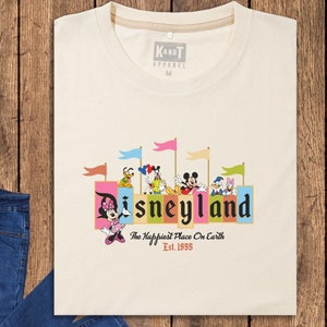 Puede incluir: Una camiseta beige con un gr&aacute;fico del logotipo de Disneyland y personajes. El texto en la camiseta dice "Disneyland, The Happiest Place On Earth, Est. 1955".