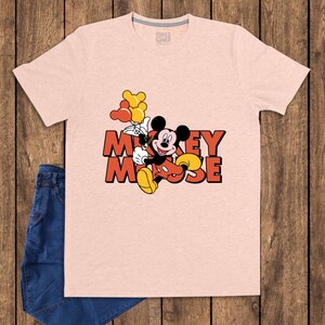 May include: A light pink t-shirt featuring a graphic of Mickey Mouse holding balloons. The text "Mickey Mouse" is in orange and red. The shirt is paired with blue jeans.