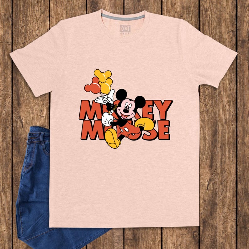 May include: A light pink t-shirt featuring a graphic of Mickey Mouse holding balloons. The text "Mickey Mouse" is in orange and red. The shirt is paired with blue jeans.