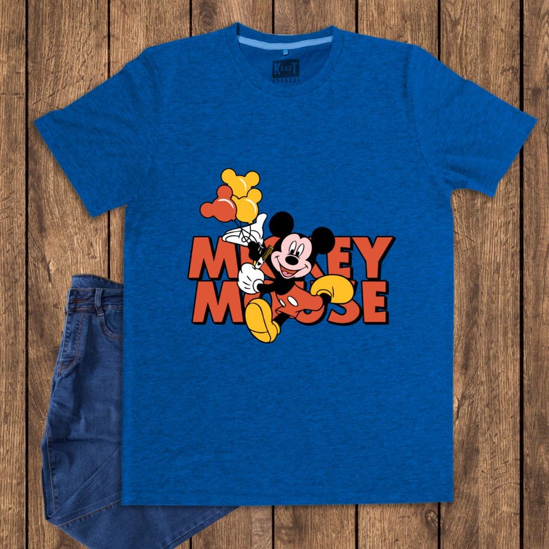 May include: A blue t-shirt featuring a graphic of Mickey Mouse holding balloons. The text "Mickey Mouse" is in orange and black. A pair of blue jeans is partially visible on a wooden background.
