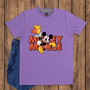 May include: A purple t-shirt featuring a graphic of Mickey Mouse holding balloons, with the words "Mickey Mouse" in orange and red. The shirt is paired with blue jeans on a wooden background.