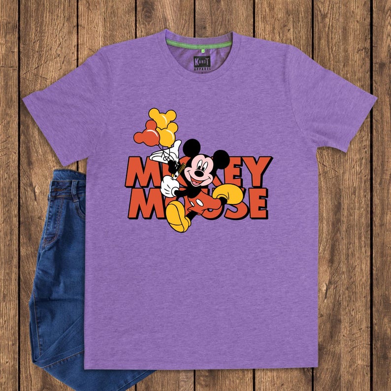 May include: A purple t-shirt featuring a graphic of Mickey Mouse holding balloons, with the words "Mickey Mouse" in orange and red. The shirt is paired with blue jeans on a wooden background.