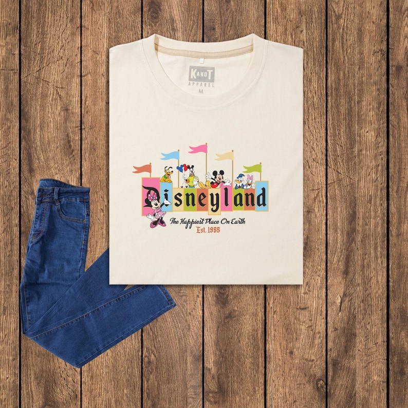 Puede incluir: Una camiseta beige con un gr&aacute;fico del logotipo de Disneyland y personajes. El texto en la camiseta dice "Disneyland, The Happiest Place On Earth, Est. 1955".