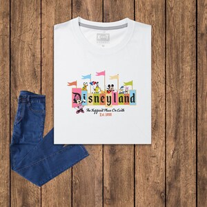 Puede incluir: Camiseta blanca con un gr&aacute;fico colorido de Mickey Mouse y otros personajes de Disney frente a un edificio con el texto "Disneyland" y "The Happiest Place On Earth" con el a&ntilde;o "Est. 1955" debajo.