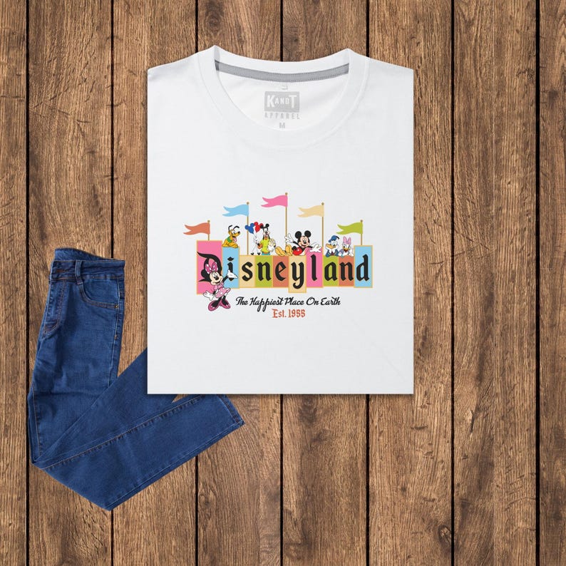 Puede incluir: Camiseta blanca con un gr&aacute;fico colorido de Mickey Mouse y otros personajes de Disney frente a un edificio con el texto "Disneyland" y "The Happiest Place On Earth" con el a&ntilde;o "Est. 1955" debajo.