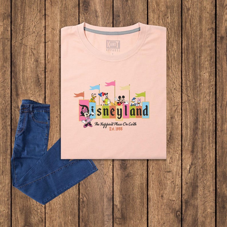 Puede incluir: Camiseta rosa con un gr&aacute;fico del logotipo de Disneyland y varios personajes de Disney. El texto dice "Disneyland, The Happiest Place On Earth, Est. 1955".