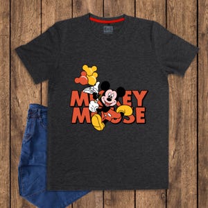May include: Dark gray t-shirt featuring Mickey Mouse holding balloons, with the words "Mickey Mouse" in orange. The shirt has a red neckline and is paired with blue jeans on a wooden surface.