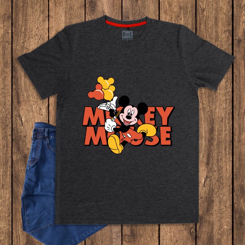 May include: Dark gray t-shirt featuring Mickey Mouse holding balloons, with the words "Mickey Mouse" in orange. The shirt has a red neckline and is paired with blue jeans on a wooden surface.