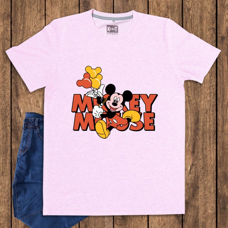May include: A light pink t-shirt featuring a graphic of Mickey Mouse holding balloons. The text "Mickey Mouse" is displayed in orange and red. The shirt is paired with blue denim jeans.