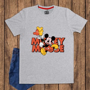 May include: A gray t-shirt featuring a graphic of Mickey Mouse holding balloons. The text "Mickey Mouse" is in orange and red. The shirt has a red collar and the brand "KaNot Apparel" is visible. Blue jeans are visible on the left.