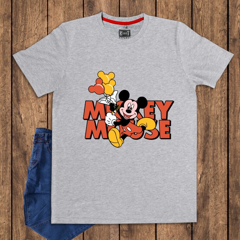 May include: A gray t-shirt featuring a graphic of Mickey Mouse holding balloons. The text "Mickey Mouse" is in orange and red. The shirt has a red collar and the brand "KaNot Apparel" is visible. Blue jeans are visible on the left.