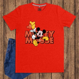 May include: A red t-shirt featuring a graphic of Mickey Mouse holding balloons, with the words "Mickey Mouse" in orange block letters. The shirt is paired with blue denim jeans. The shirt is made of a soft material.