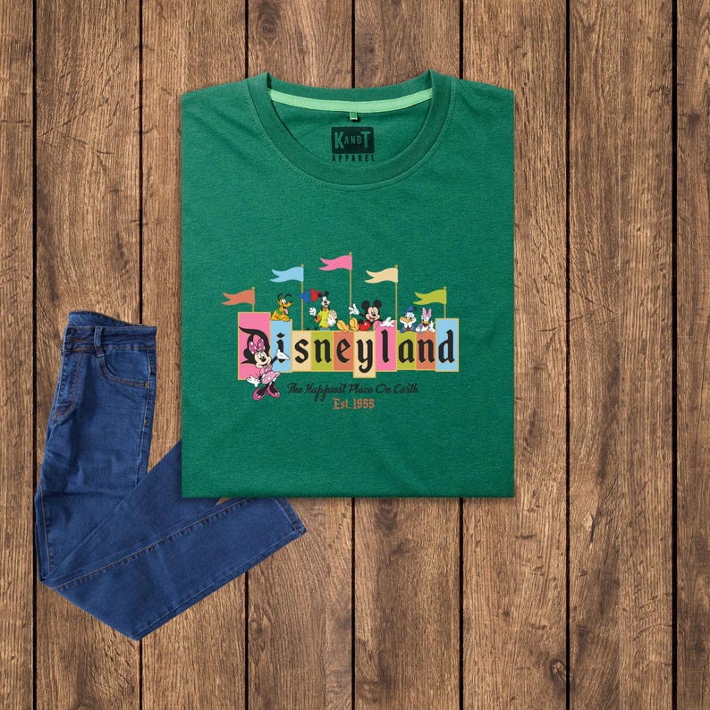 Puede incluir: Camiseta verde con un gr&aacute;fico de Mickey Mouse y otros personajes de Disney frente a un cartel de Disneyland colorido. El texto en el cartel dice "Disneyland" y "The Happiest Place On Earth" con el a&ntilde;o "Est. 1955" debajo.