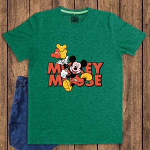May include: Green t-shirt featuring a graphic of Mickey Mouse holding balloons, with the text "Mickey Mouse" in orange and yellow. The shirt is paired with blue denim jeans. The shirt has a crew neck and short sleeves.