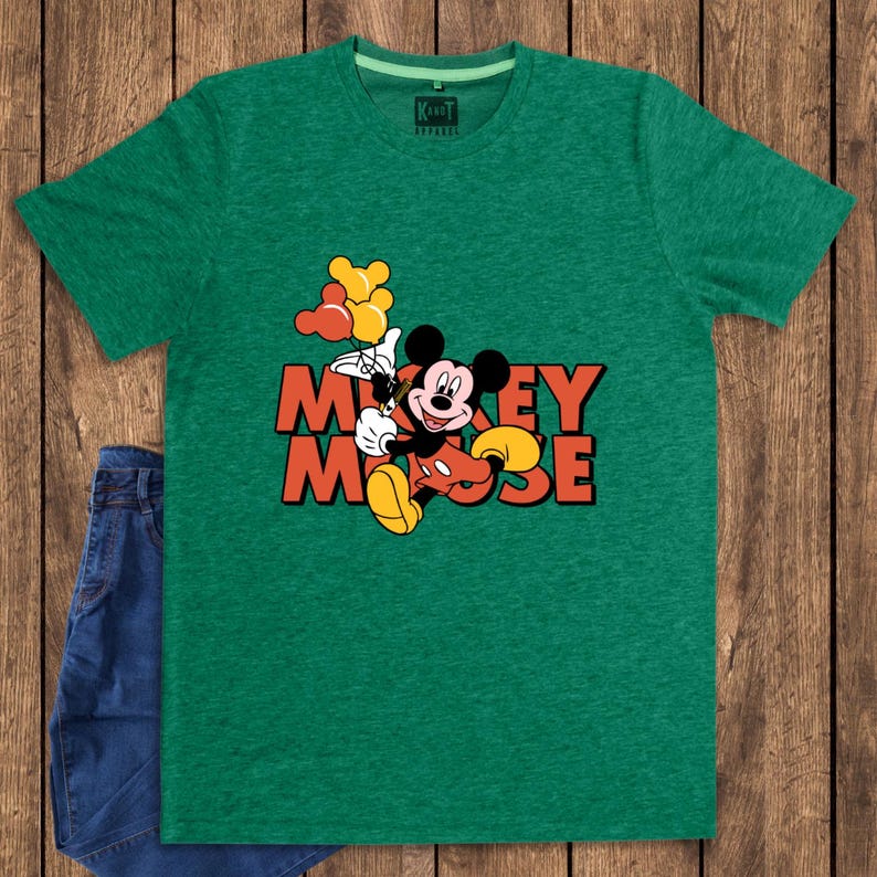 May include: Green t-shirt featuring a graphic of Mickey Mouse holding balloons, with the text "Mickey Mouse" in orange and yellow. The shirt is paired with blue denim jeans. The shirt has a crew neck and short sleeves.
