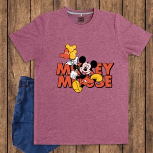 May include: Maroon t-shirt featuring a graphic of Mickey Mouse holding balloons, with the words "Mickey Mouse" in orange. The shirt is paired with blue denim jeans. The shirt has a crew neck and short sleeves.
