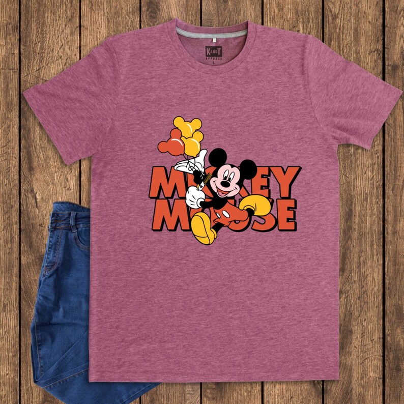 May include: Maroon t-shirt featuring a graphic of Mickey Mouse holding balloons, with the words "Mickey Mouse" in orange. The shirt is paired with blue denim jeans. The shirt has a crew neck and short sleeves.