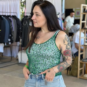 May include: A green sequined tank top with a spaghetti strap design. The top is paired with light blue denim jeans. The image is taken in a retail setting, with clothing racks and other shoppers in the background.