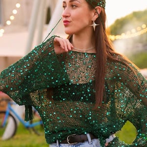Pode incluir: Um top verde de tecido aberto com mangas compridas e lantejoulas espalhadas. O top é combinado com jeans de lavagem clara e um cinto preto. Um gorro verde a condizer e brincos em forma de estrela completam o look. O traje é adequado para um ambiente casual ou festivo.