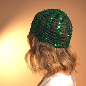 May include: A dark green crochet cap featuring an open-knit lattice pattern adorned with small, reflective green sequins throughout the structure. The headwear is fitted, covering the crown of the head with a textured, yarn-based construction.