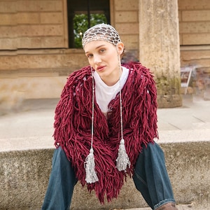 May include: A burgundy, shaggy knit jacket with long fringe detail. The jacket is worn over a white t-shirt and paired with blue jeans. Silver beaded tassels hang from the jacket. A silver headpiece is also worn.
