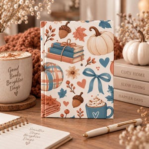 Puede incluir: Un diario de tapa dura con una cubierta blanca que presenta un patrón repetitivo de calabazas, libros azules atados con cuerda, bellotas, hojas de otoño, lazos azules, corazones y una taza azul de chocolate caliente con crema batida.