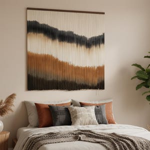 Könnte beinhalten: Ein Schlafzimmer mit einem Bett, das mit einer hellen Bettdecke und einer strukturierten Decke ausgestattet ist. Dekorative Kissen in Grau- und Rosttönen sind auf dem Bett angeordnet. Über dem Bett hängt ein großer Wandbehang mit einem Landschaftsdesign in neutralen Tönen.