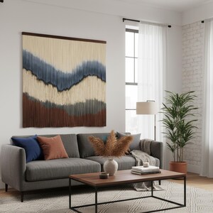Op de afbeelding: Een moderne woonkamer met een grijze bank, blauwe en oranje kussens en een houten salontafel. Boven de bank hangt een groot wandkleed met lagen in blauw, bruin en beige. De kamer heeft een neutraal kleurenpalet en een patroonkleed.