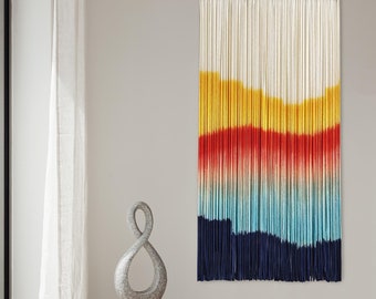 Puede incluir: Un tapiz de pared colorido hecho de hilo con un degradado de amarillo, naranja, rojo, azul y azul marino. El tapiz está hecho de hilos individuales que se tejen juntos para crear un efecto texturizado.