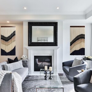 Peut inclure: Deux tentures murales en macram&eacute; noir et blanc avec un motif ondul&eacute; sont accroch&eacute;es &agrave; un mur blanc au-dessus d'un canap&eacute; gris et de deux fauteuils gris. Un miroir encadr&eacute; noir est suspendu au-dessus d'une chemin&eacute;e blanche avec un &eacute;cran de chemin&eacute;e noir.