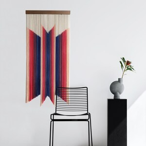 May include: A colorful macrame wall hanging with a geometric design in shades of red, blue, and white. The hanging is made of yarn and is attached to a wooden dowel.