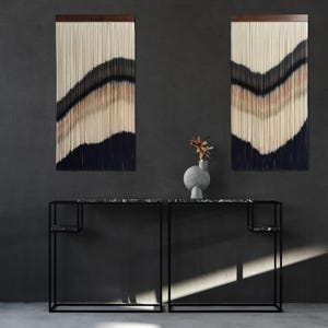 Peut inclure: Deux consoles en m&eacute;tal noir avec des plateaux en marbre noir et une seule &eacute;tag&egrave;re. Les consoles sont plac&eacute;es devant un mur gris avec deux tentures murales en macram&eacute; dans un sch&eacute;ma de couleurs beige et bleu. Un vase gris avec un arrangement de fleurs s&eacute;ch&eacute;es repose sur les consoles.