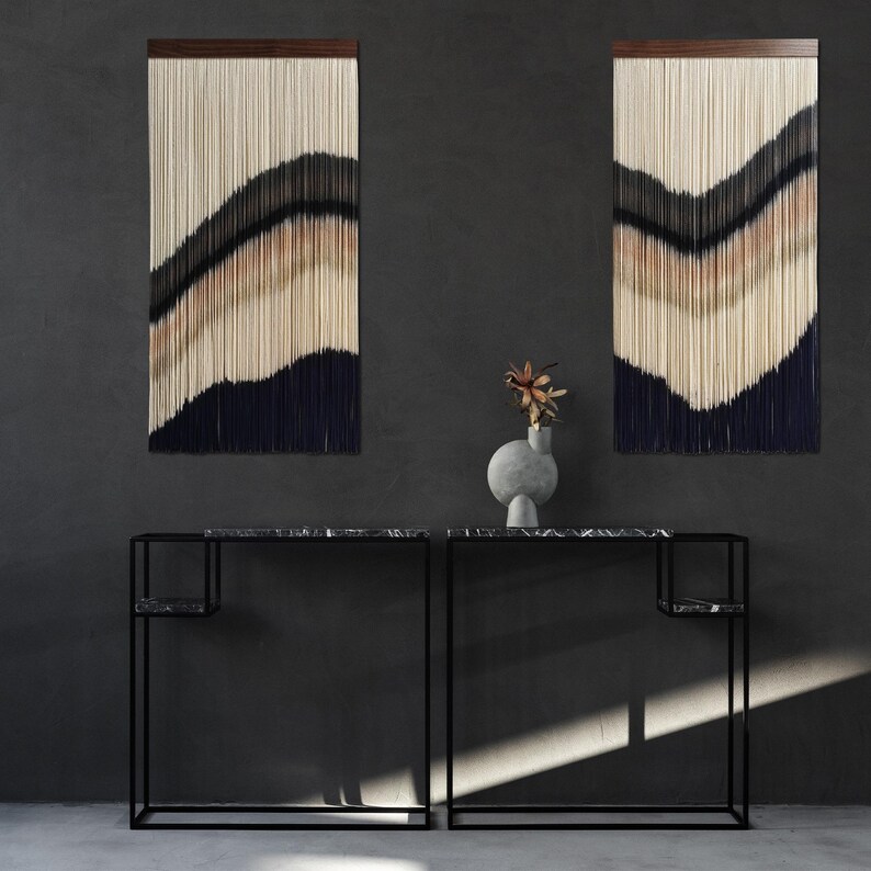 Peut inclure: Deux consoles en m&eacute;tal noir avec des plateaux en marbre noir et une seule &eacute;tag&egrave;re. Les consoles sont plac&eacute;es devant un mur gris avec deux tentures murales en macram&eacute; dans un sch&eacute;ma de couleurs beige et bleu. Un vase gris avec un arrangement de fleurs s&eacute;ch&eacute;es repose sur les consoles.