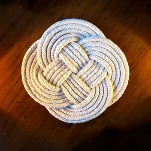 May include: A white rope coaster with a knotted design. The coaster is round and has a woven texture.